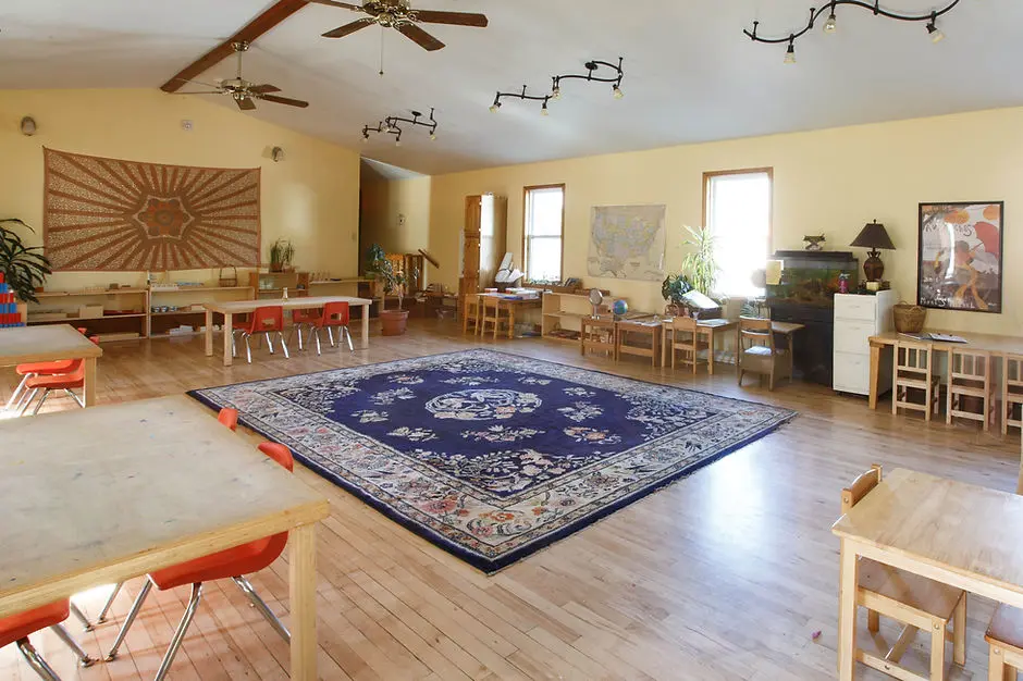 A large, open classroom space features child-sized furniture and a central patterned rug.