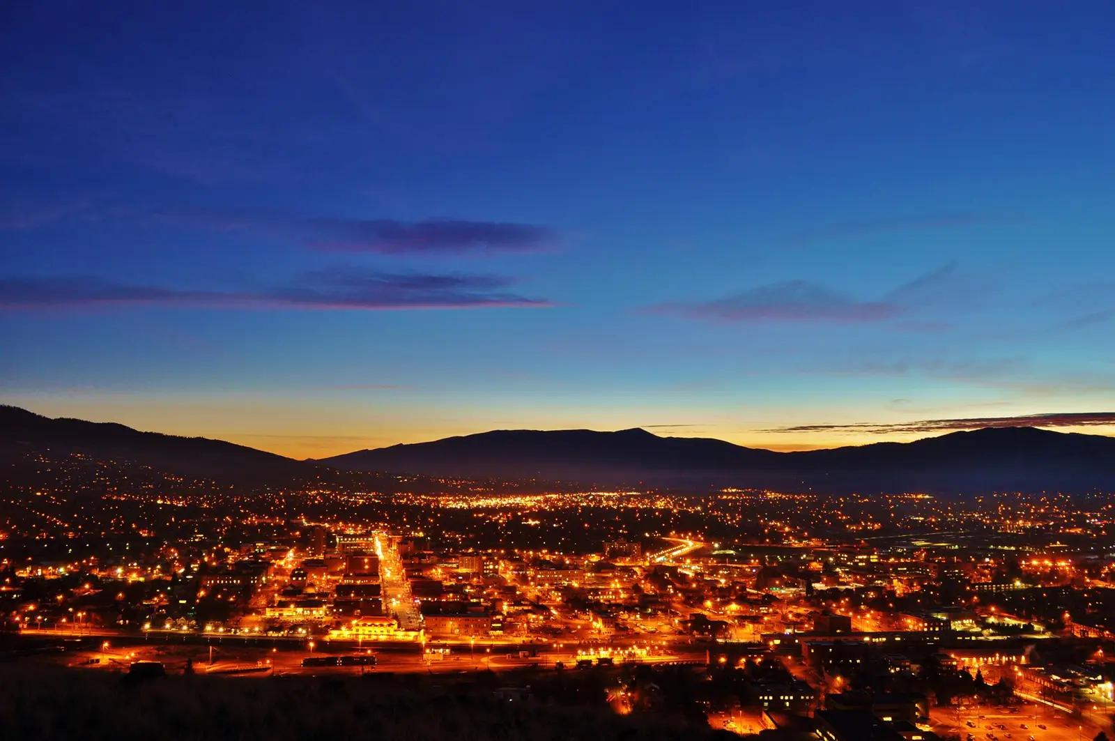 A night view of a town illuminated by city lights with mountains in the background under a twilight sky.