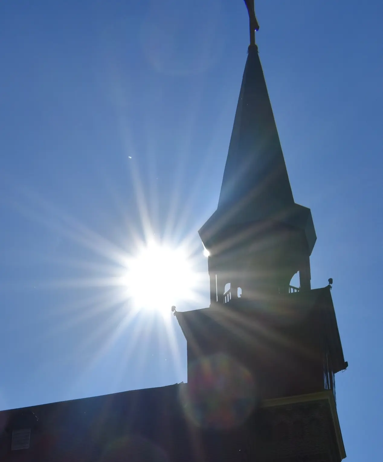 The silhouette of a church steeple is backlit by the sun against a clear blue sky.