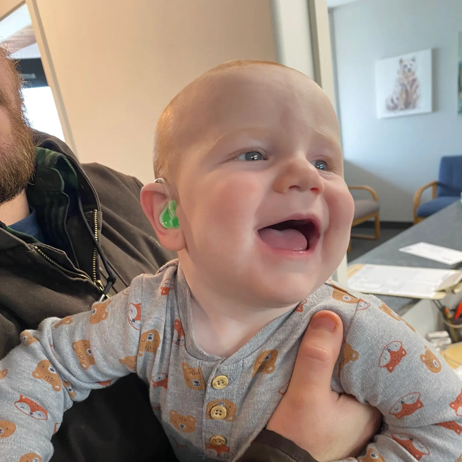 A smiling baby wearing a hearing aid is held by a person.