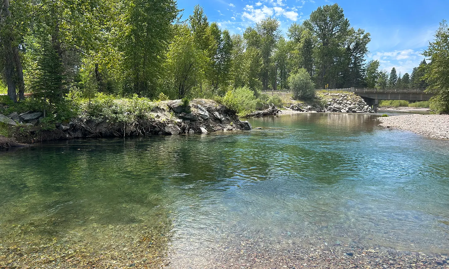 A clear, shallow river flows past a rocky bank under a bright blue sky.