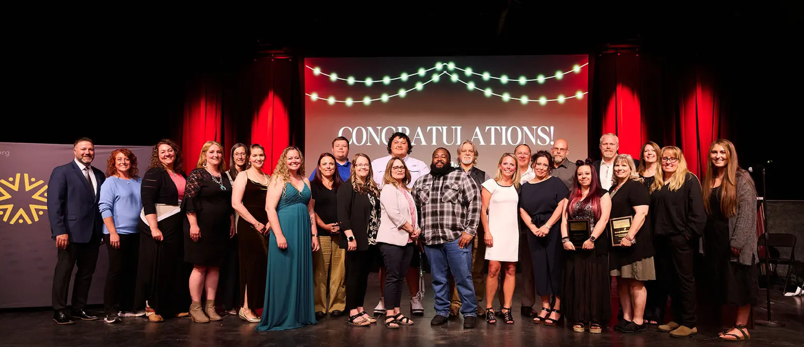 A large group of adults stands on a stage in front of a screen displaying the text CONGRATULATIONS!