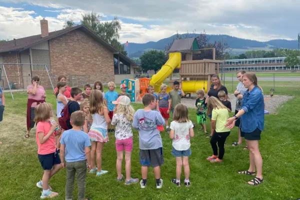 A group of children and adults are gathered on a grassy field in front of a playground structure.