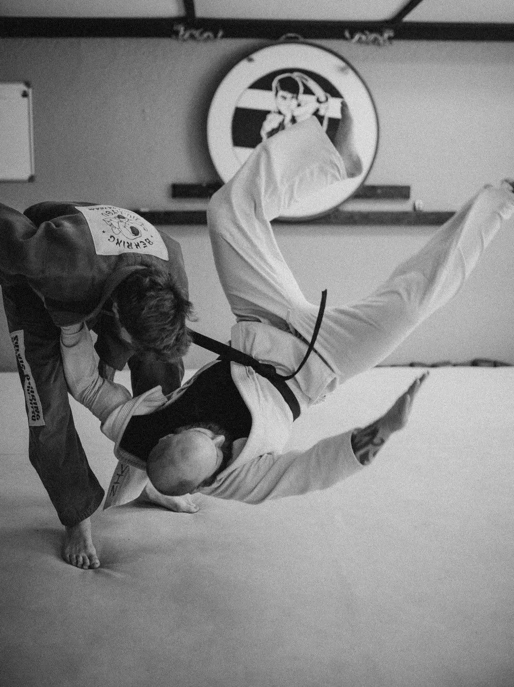 Two adults in martial arts uniforms are practicing a throw in a studio.
