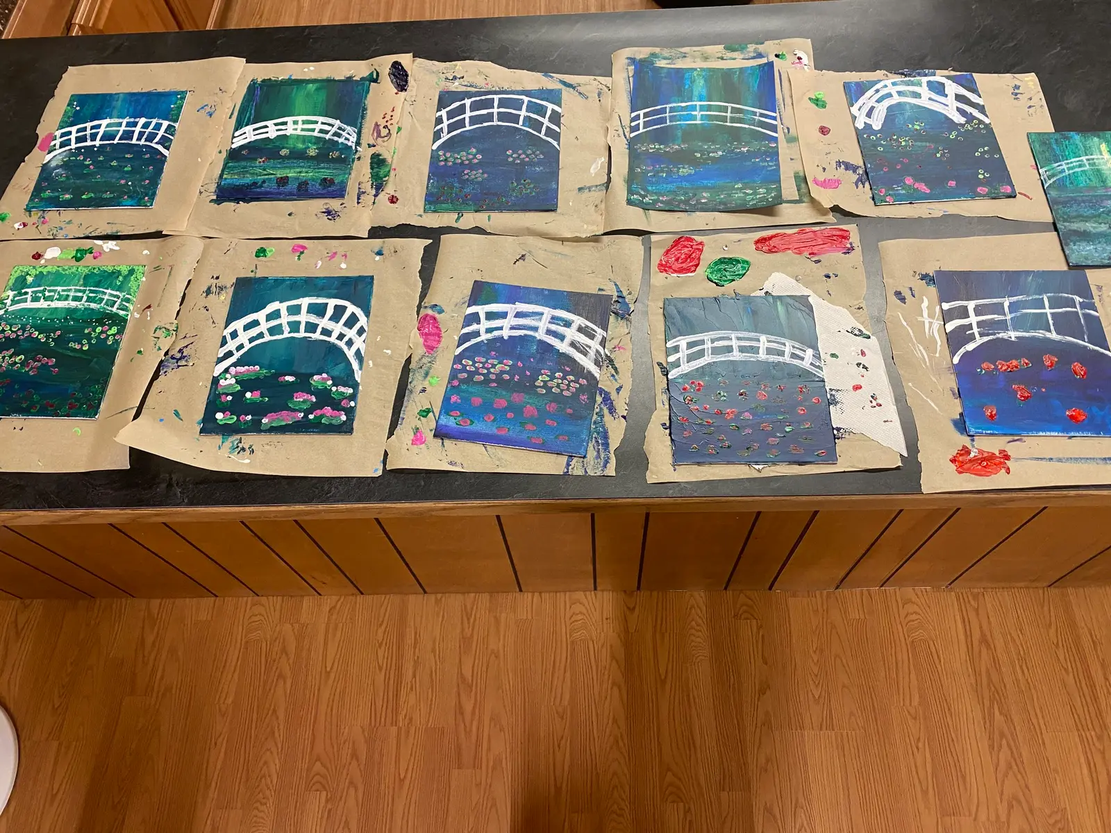 Several finished paintings of bridges over water are laid out on a table.