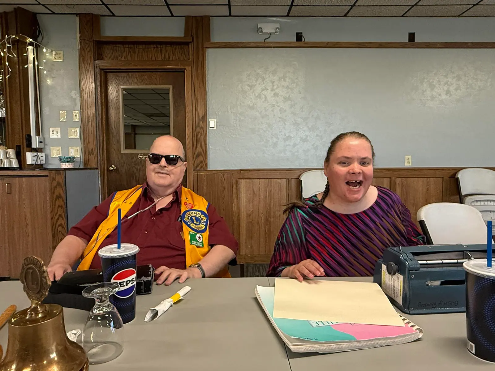 Two adults sit together at a table with drinks and a braille writer.
