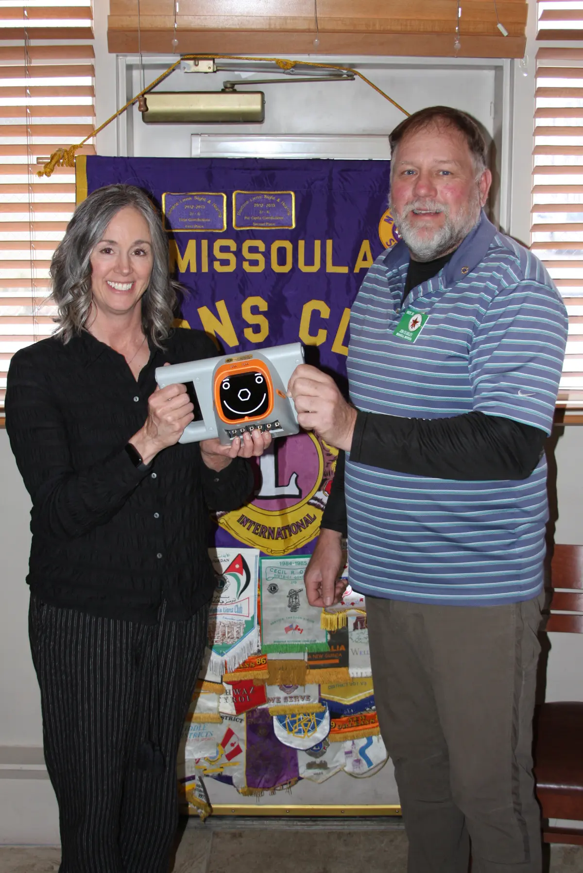 A woman and a man hold a small device with a smiling face screen in front of a club banner.