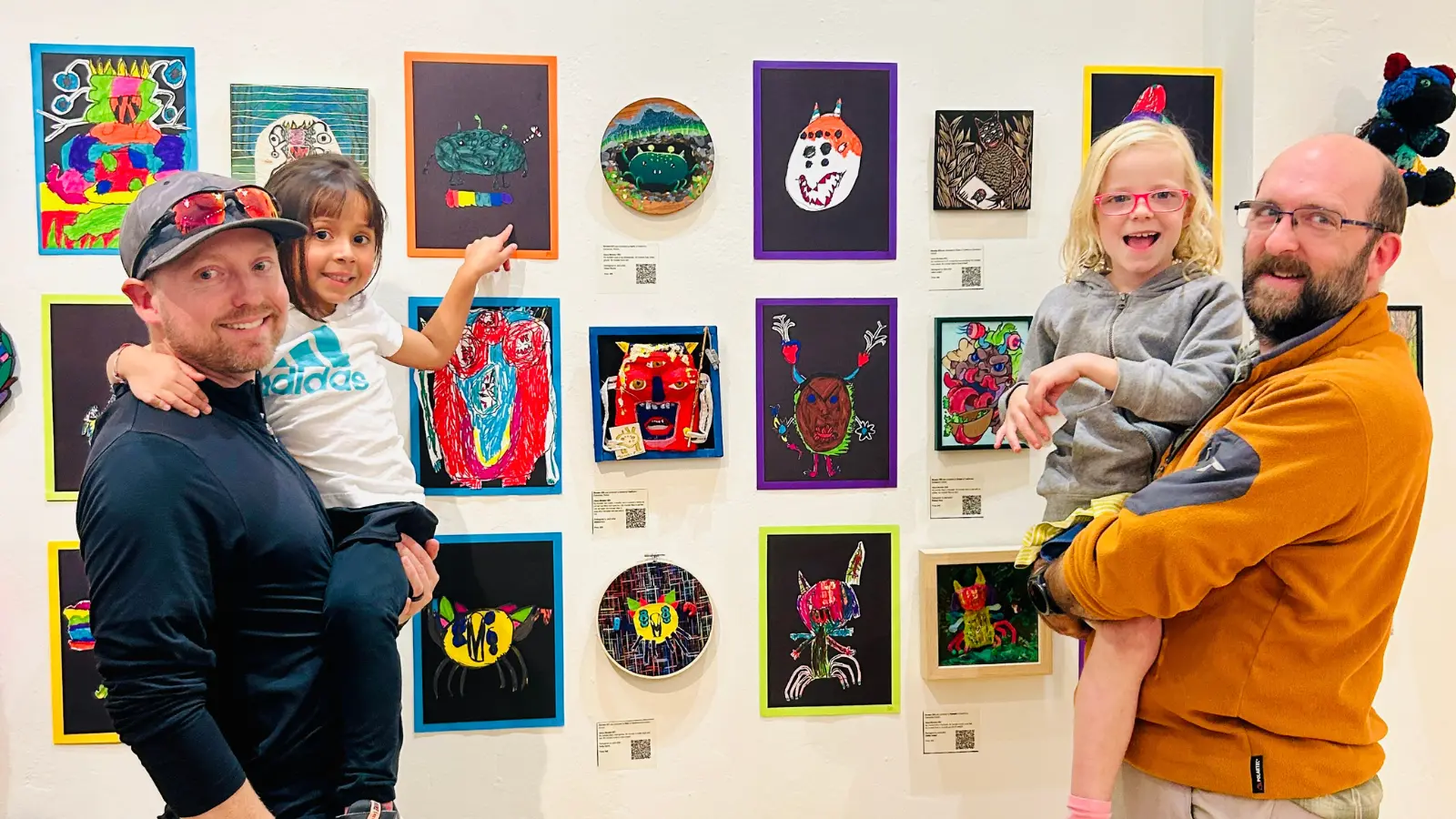 Two adults holding young children in front of a wall decorated with various children's drawings.