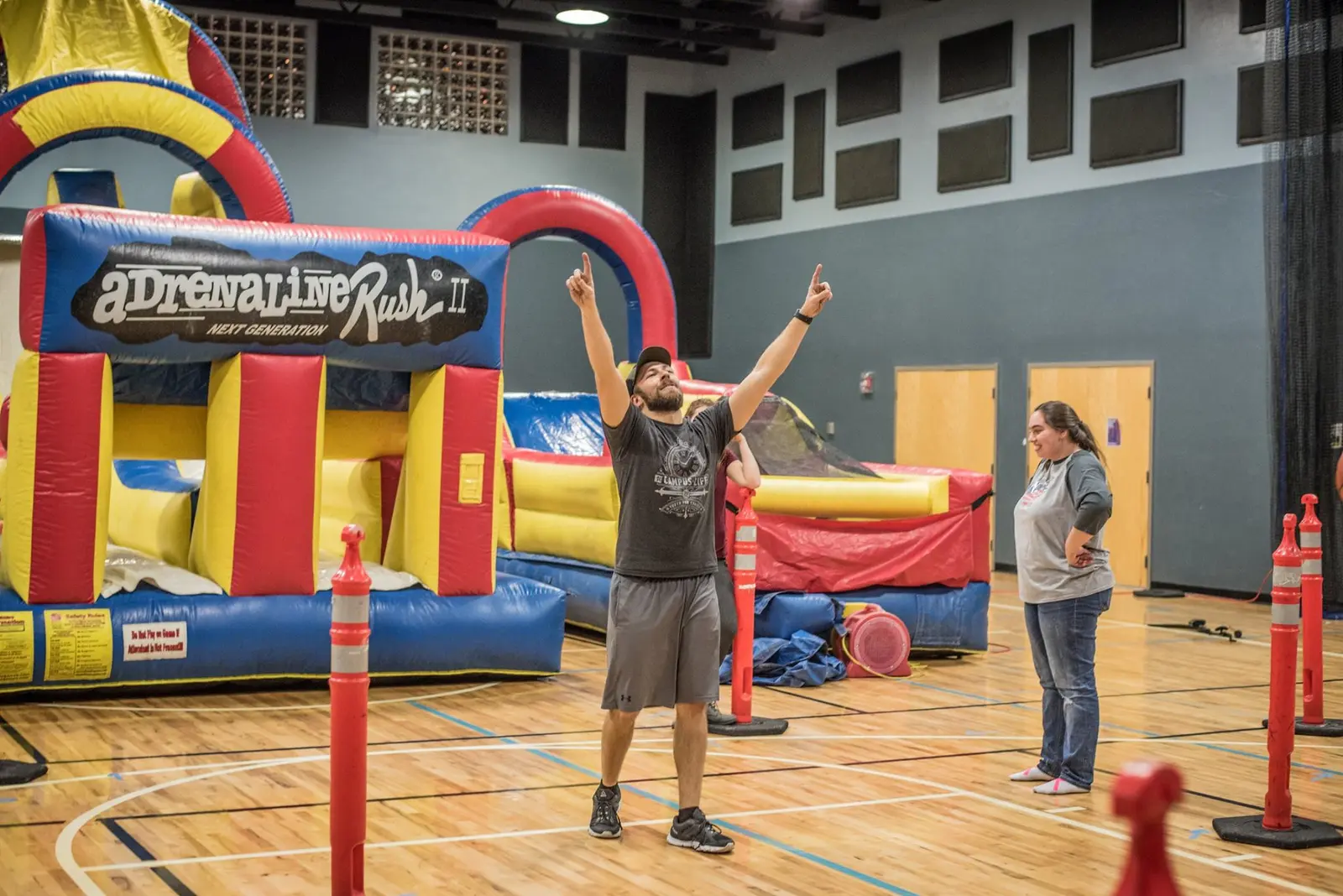 A man stands with arms raised in front of an inflatable obstacle course in a gymnasium.
