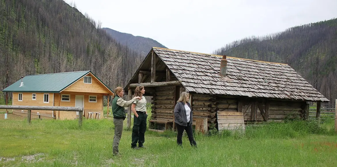 Three people stand in a field with two cabins near a burnt forest.