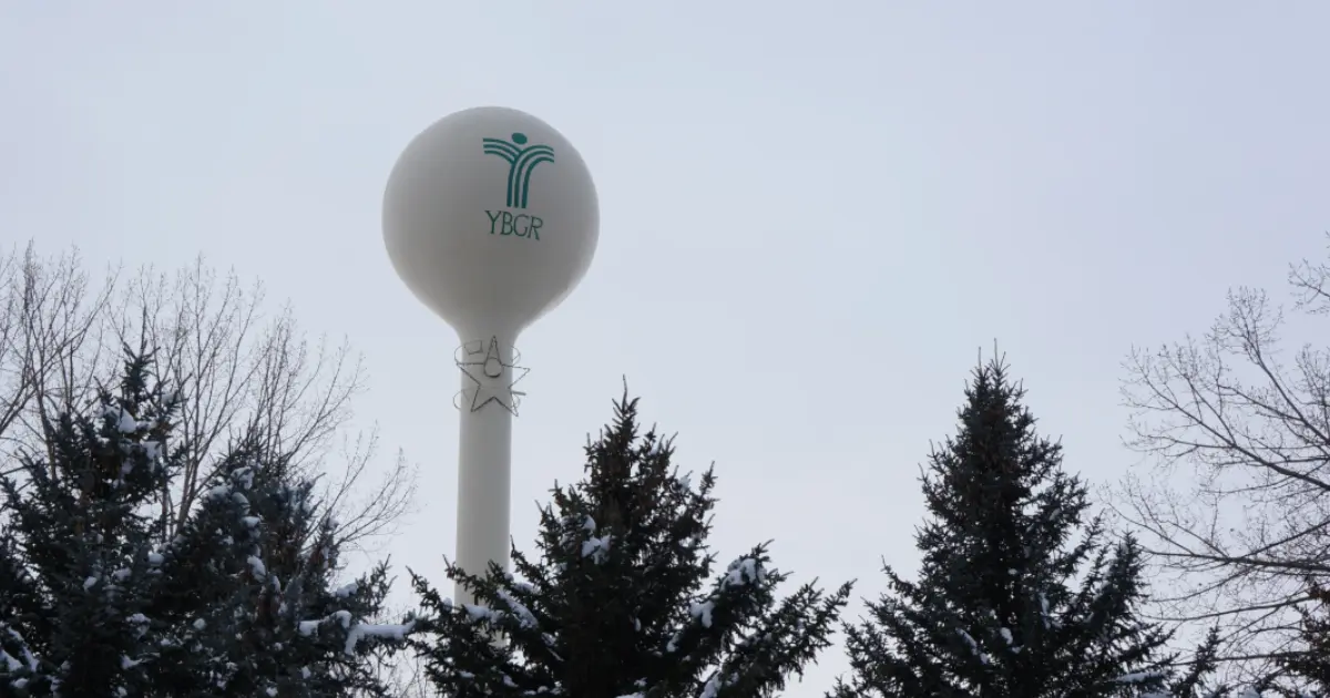 A water tower with a logo stands among snow-dusted evergreen trees.