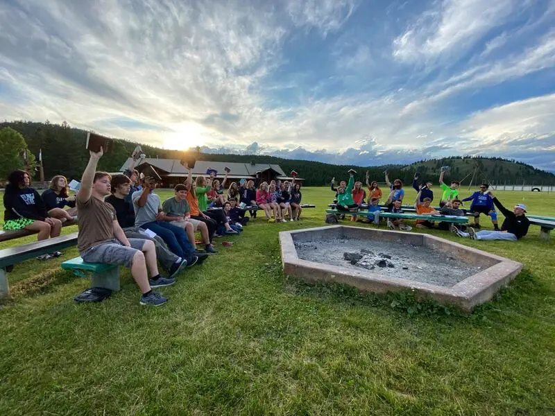A group of young people sits on benches outdoors near a fire pit while holding up books.