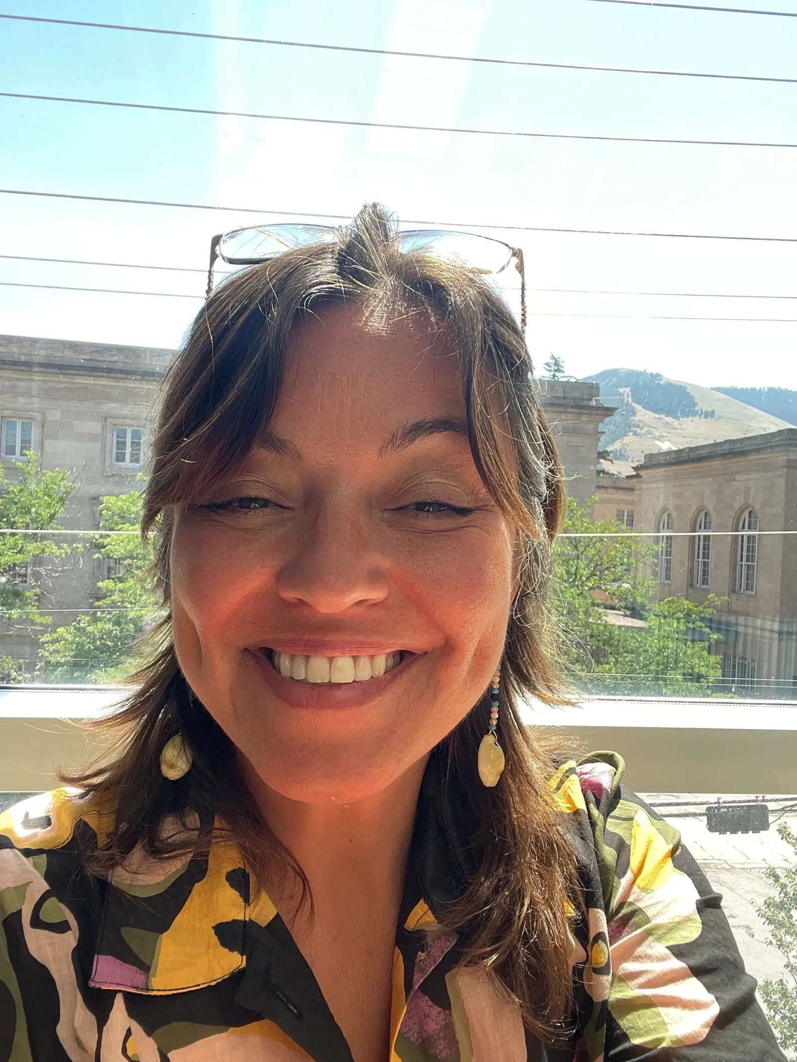 A smiling woman wears glasses on her head and earrings in front of a window overlooking a building and mountain.
