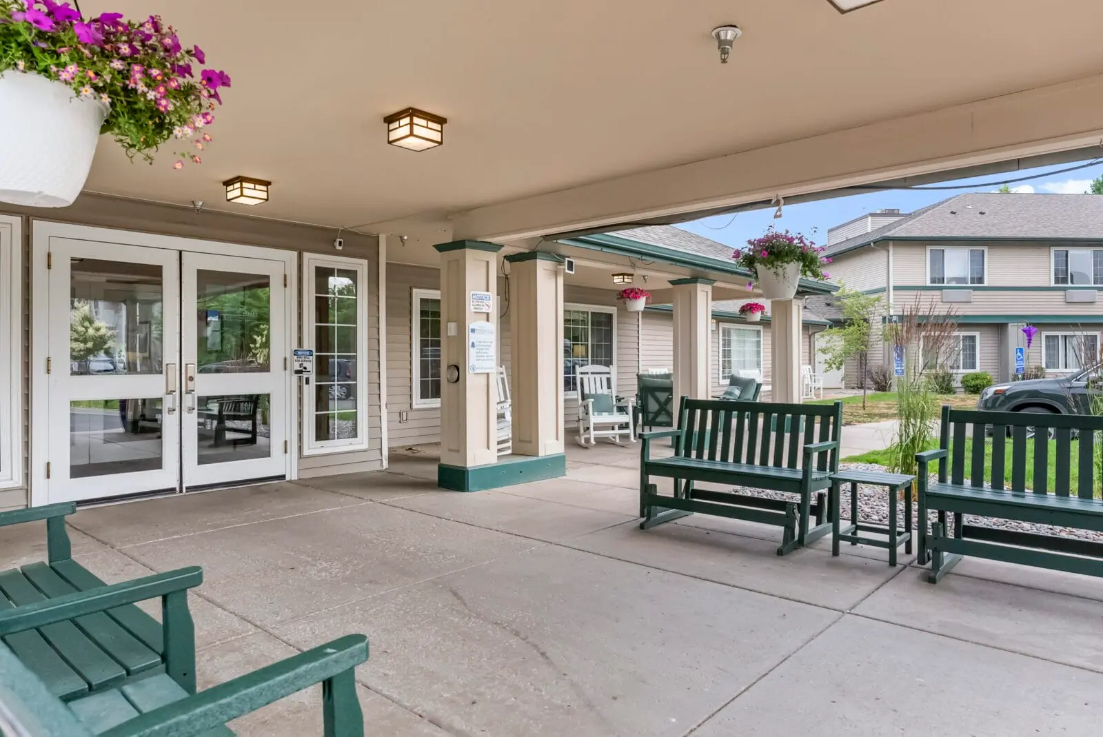 A shaded outdoor patio area of a building features green benches, hanging flower baskets, and a glass-paned entrance.
